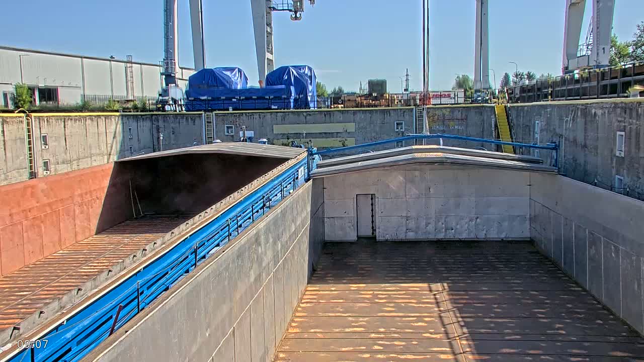 The interior of a large, empty cargo barge is shown, with concrete walls and a blue metal railing, under a sunny sky.