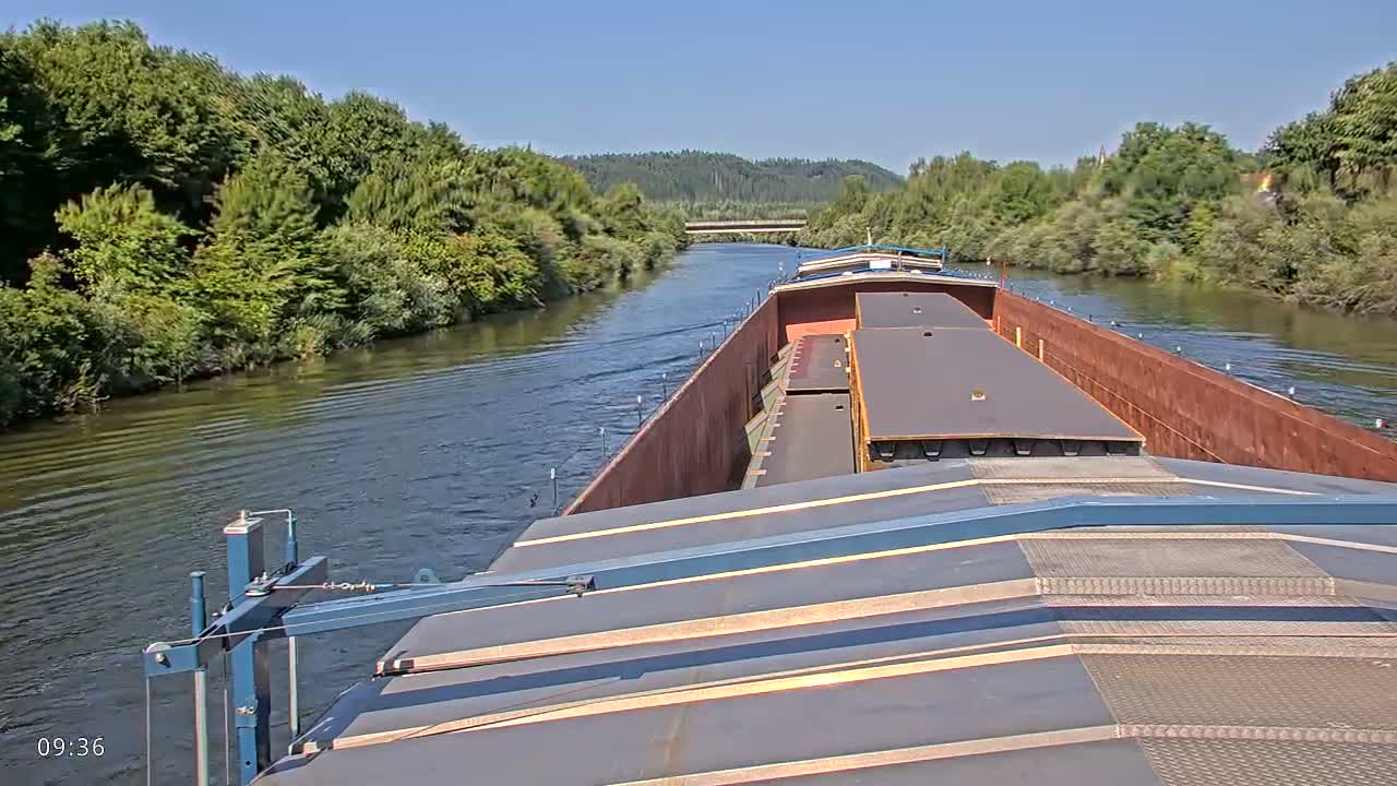 A barge travels down a river on a sunny day, surrounded by lush green vegetation.