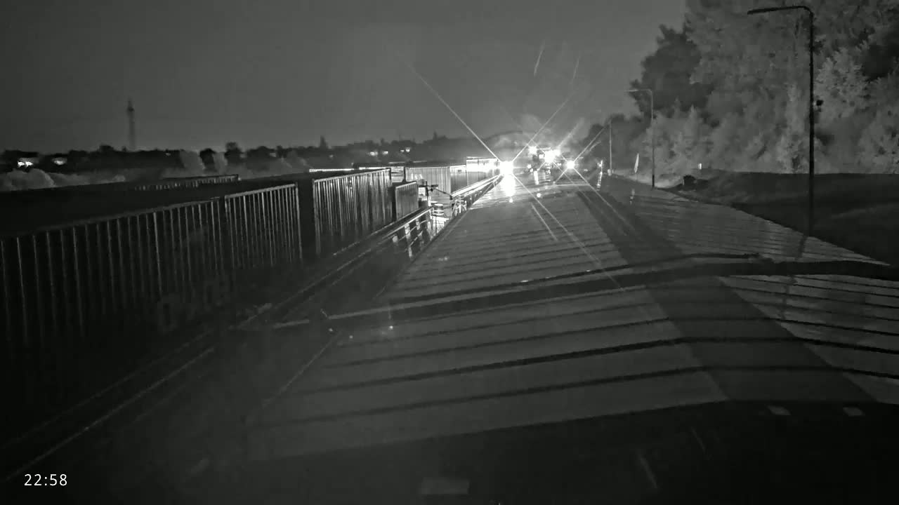 A nighttime, rainy view from atop a train car shows a long line of other train cars ahead, lit by their headlights.