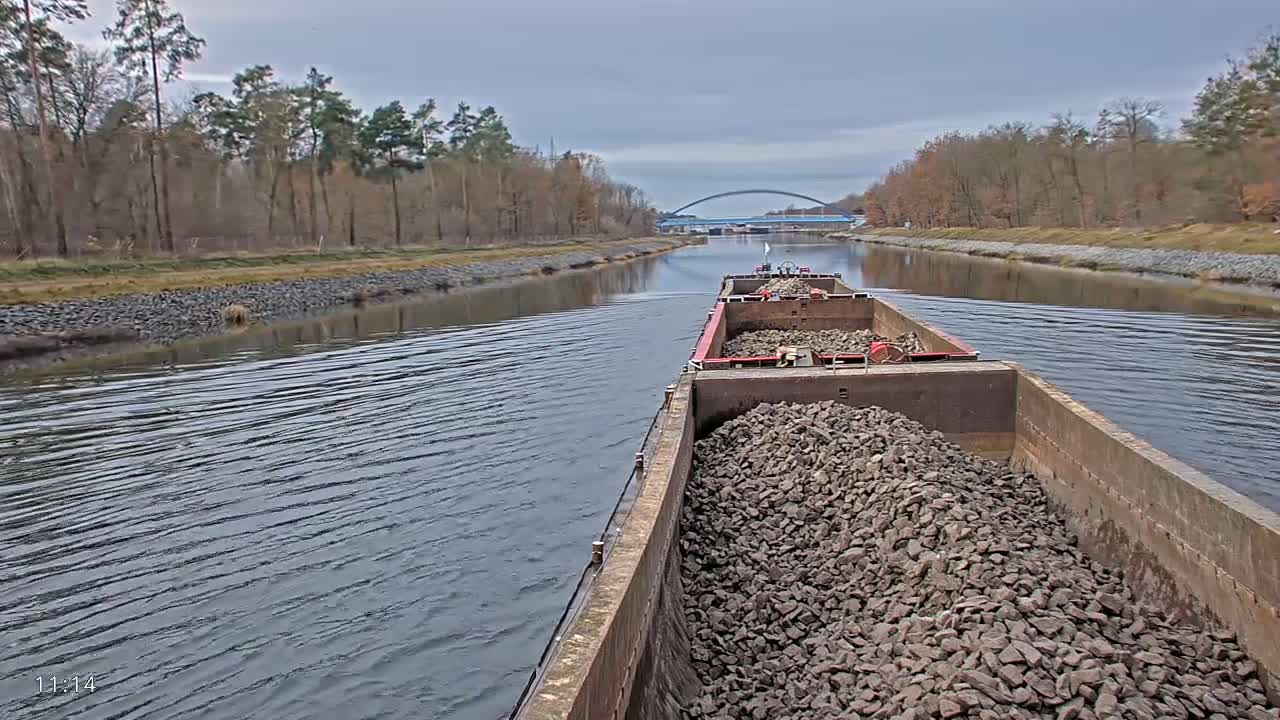 A cargo barge laden with rocks navigates a calm canal flanked by sparse trees, heading towards a distant blue arch bridge under an overcast sky.