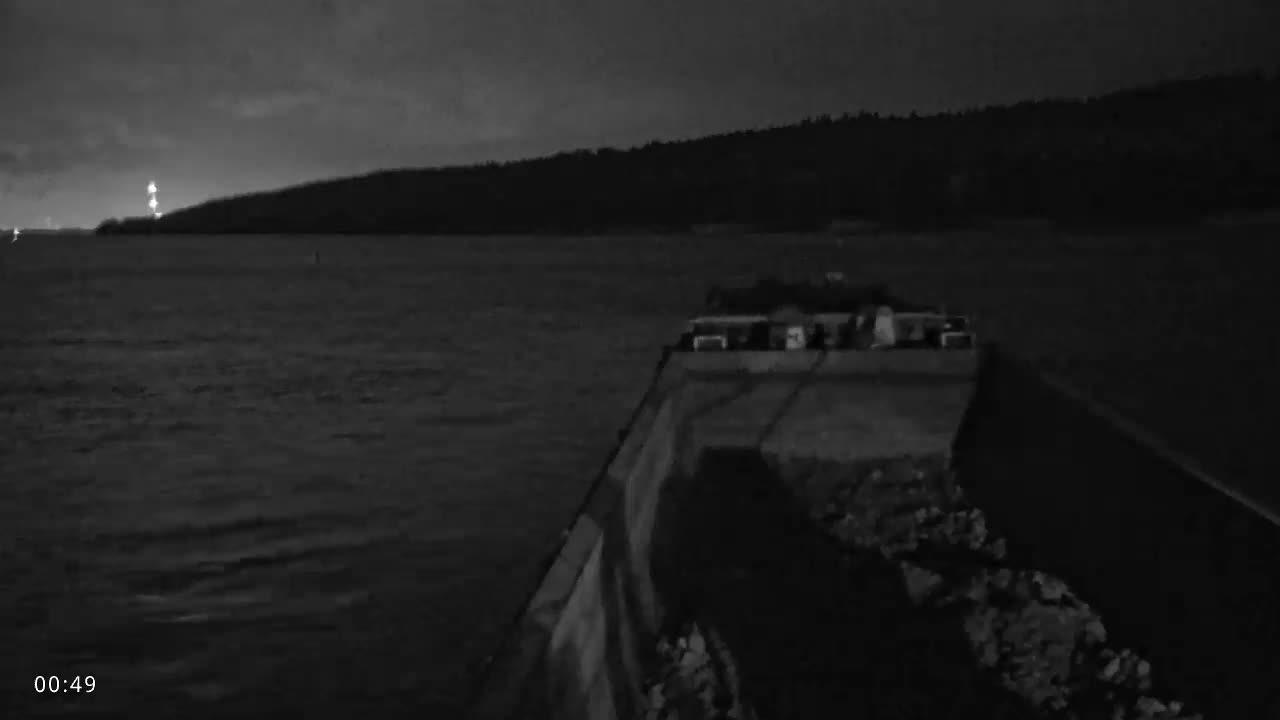 A monochromatic view on a cloudy night shows a calm body of water with a distant lighthouse on a wooded coastline, seen from a concrete breakwater in the foreground.