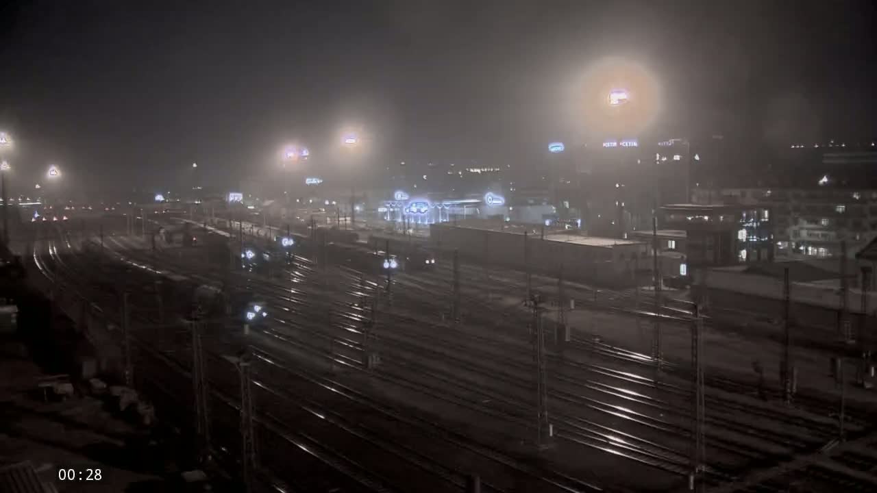 A foggy night scene shows a train yard with numerous tracks and trains, illuminated by lights, alongside buildings in the background.
