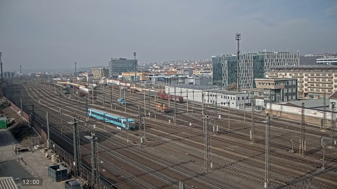 A vast railway yard with numerous train cars and tracks, under a hazy sky, is situated next to a city featuring modern and older buildings.