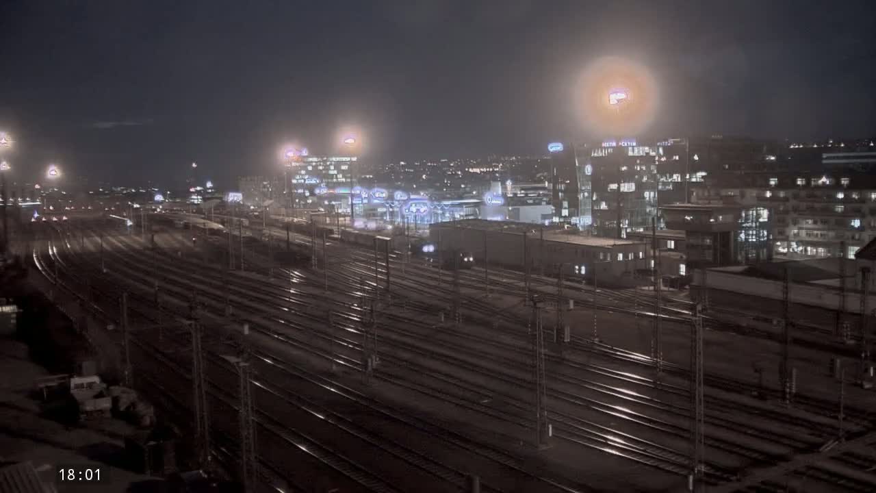 A nighttime view of a large railway yard with many tracks extending into a city featuring illuminated buildings under a hazy sky.