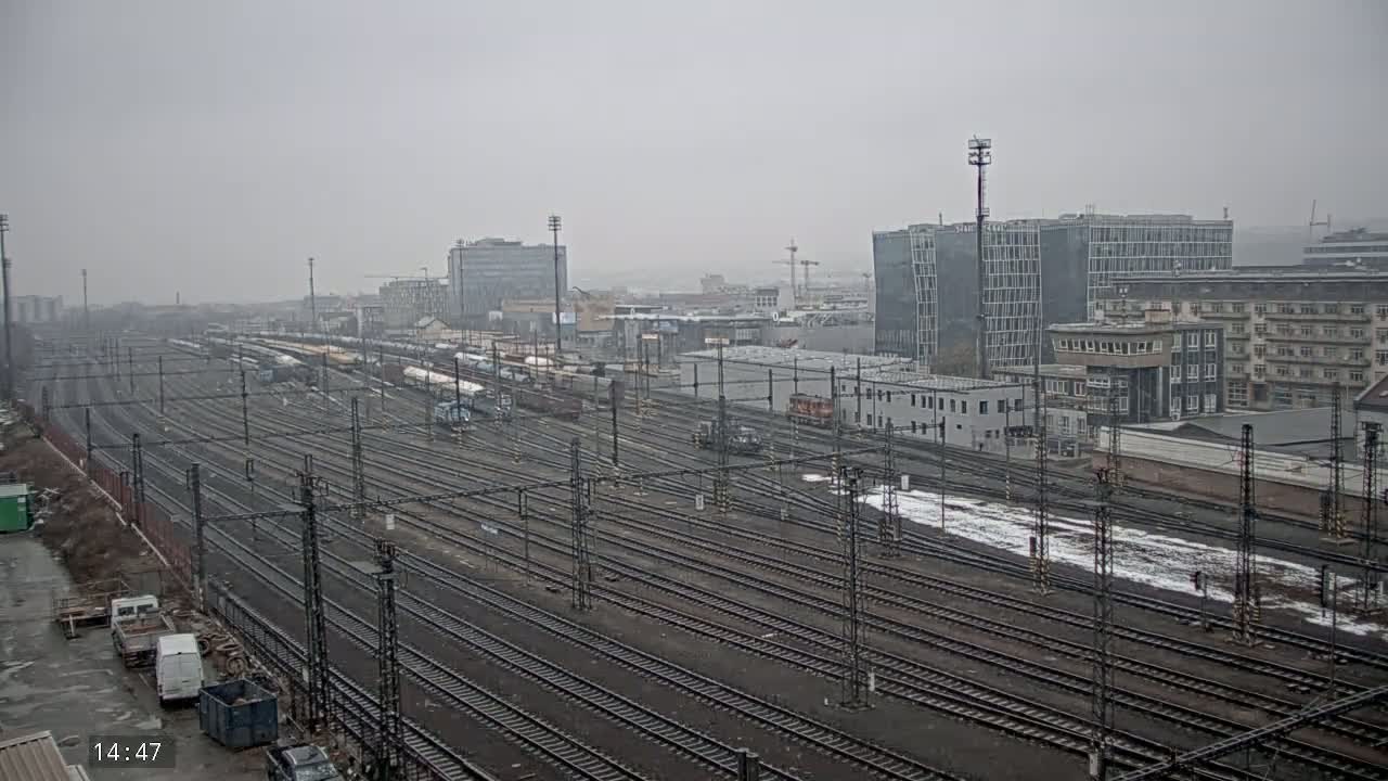 A sprawling railway yard with numerous tracks and parked trains is visible under a hazy, overcast sky, with a few buildings in the background.