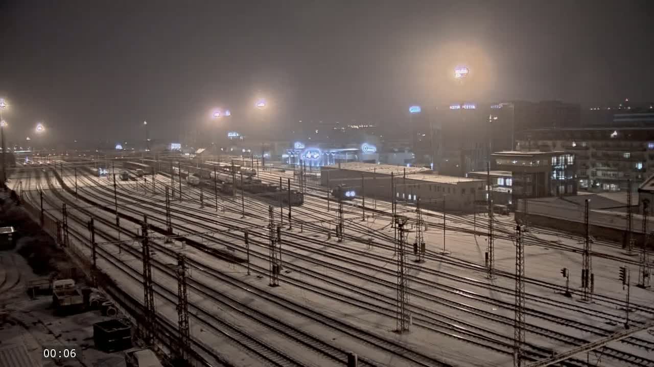 A snow-covered train yard at night, illuminated by numerous lights, with several buildings in the background.