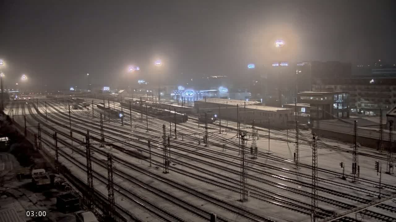 A snow-covered train yard at night, illuminated by numerous lights, with trains parked on the tracks and buildings in the background.