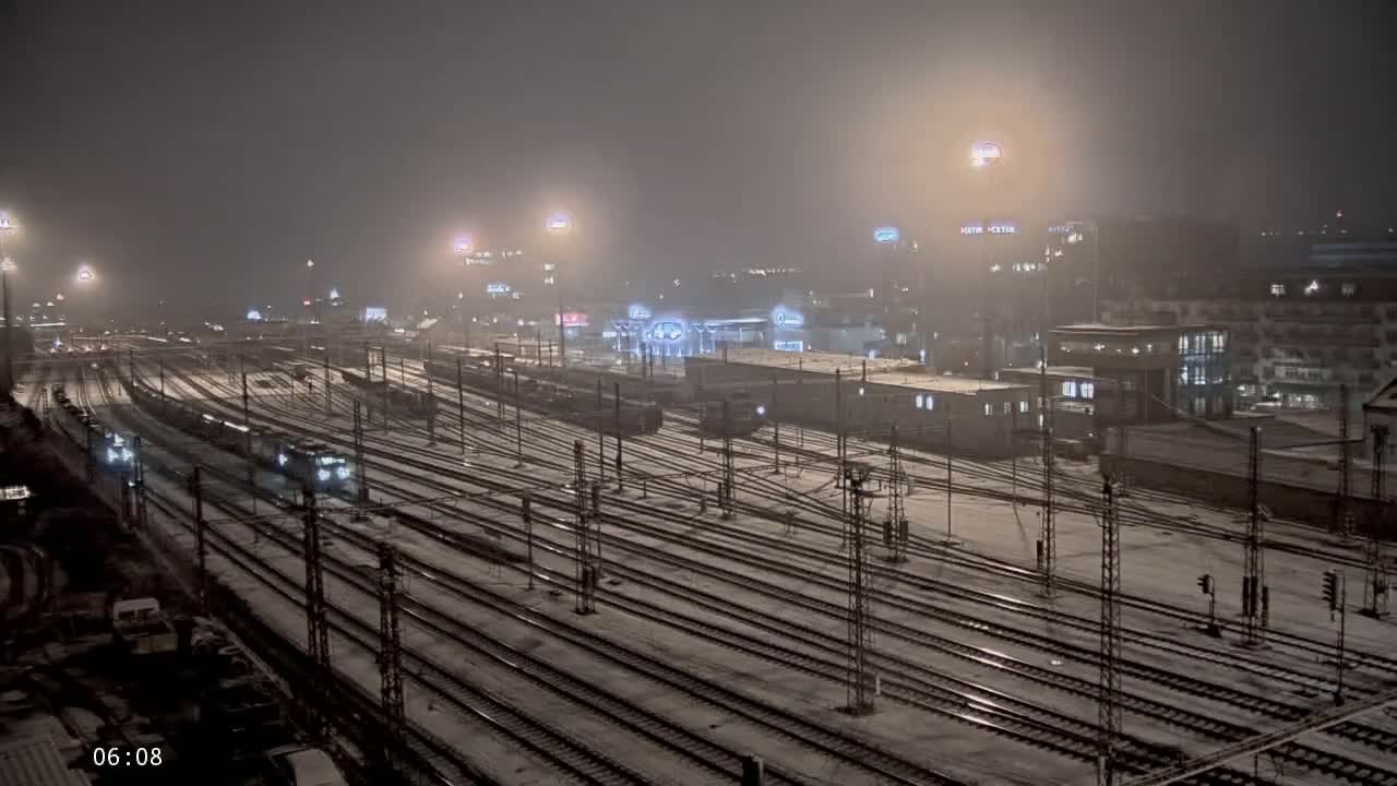 A snowy night scene at a train station shows numerous train tracks covered in snow with several trains visible under dim lighting.