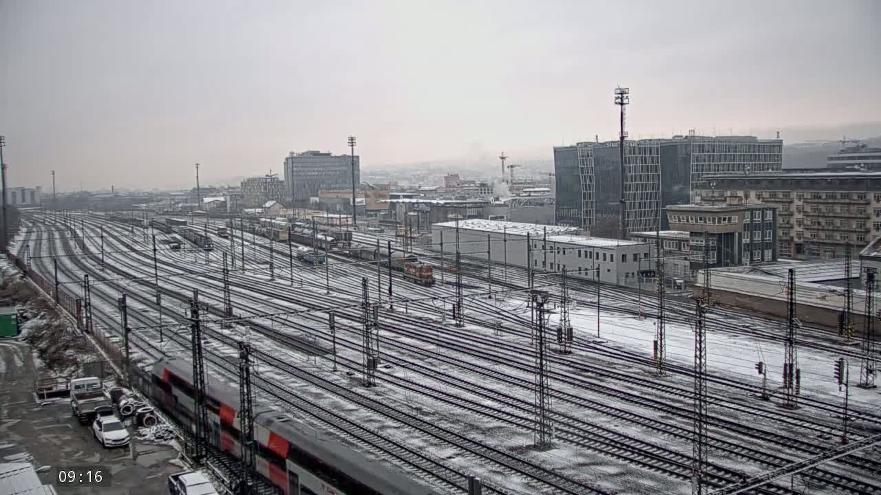 A snow-covered railway yard with numerous train tracks extends toward a city skyline under an overcast sky.