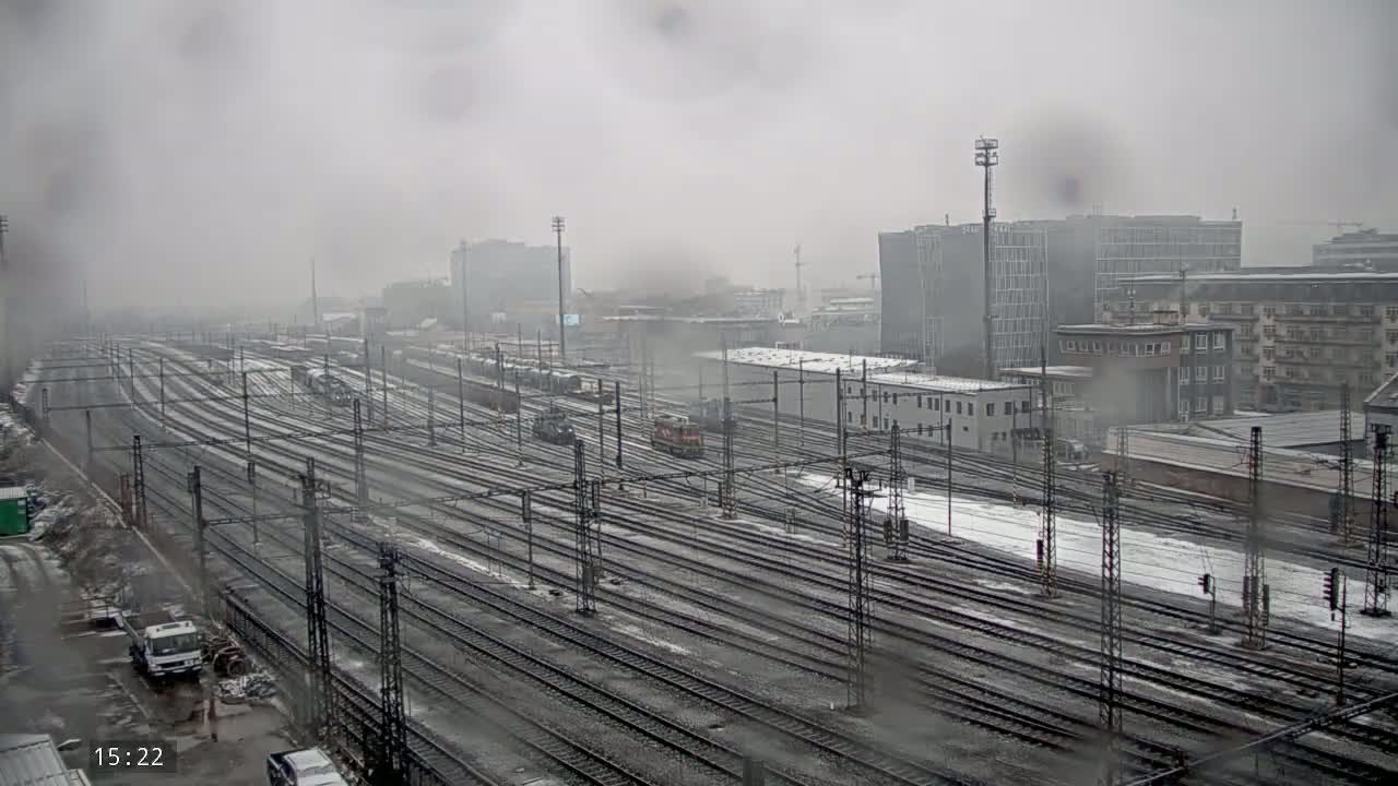 A snowy, overcast day reveals a vast railway yard with numerous train tracks, some trains, and buildings in the background.