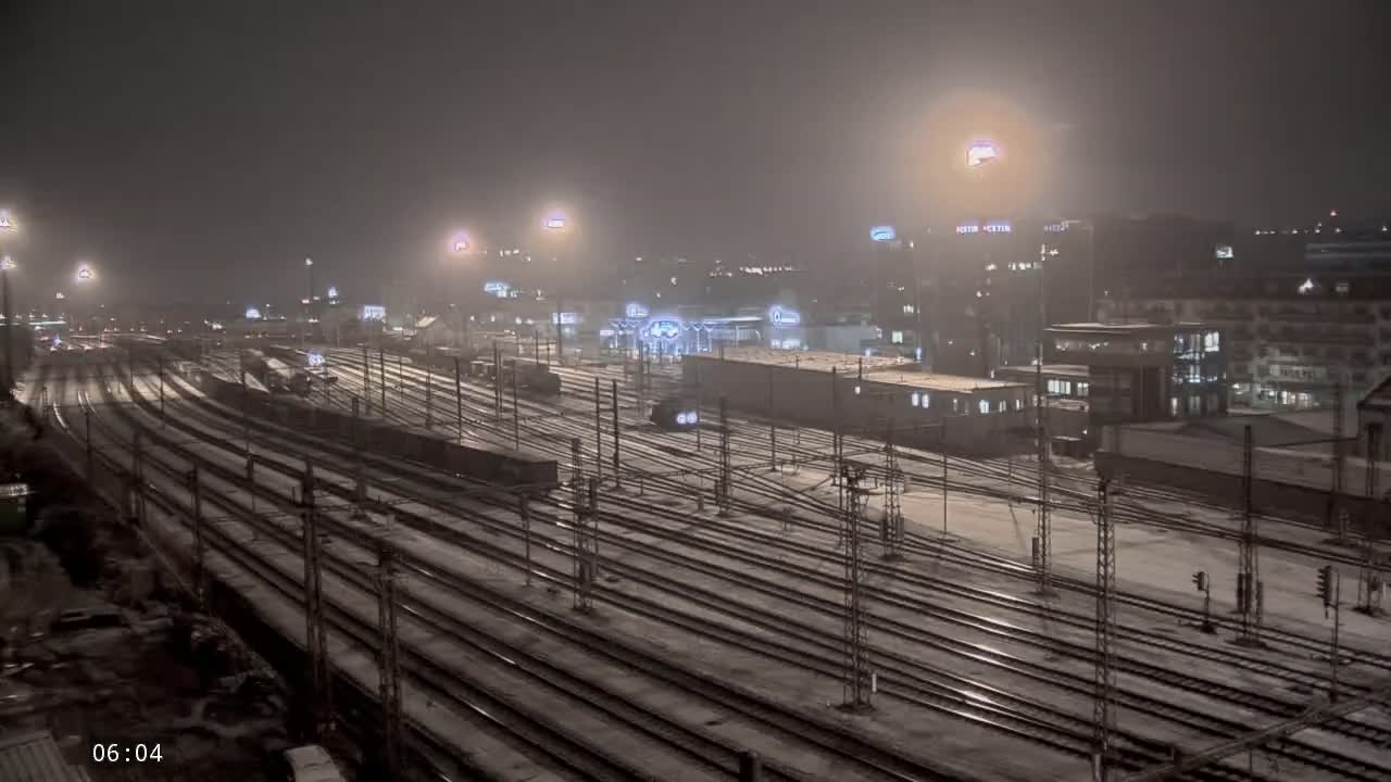 A snow-covered railway yard at night, illuminated by numerous lights, with buildings in the background.