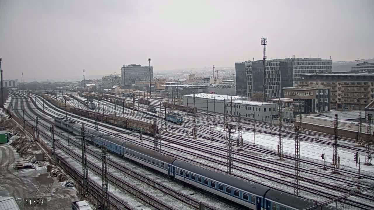 A snow-covered railway yard with numerous train tracks and several passenger and freight trains, situated near a city with modern and older buildings.
