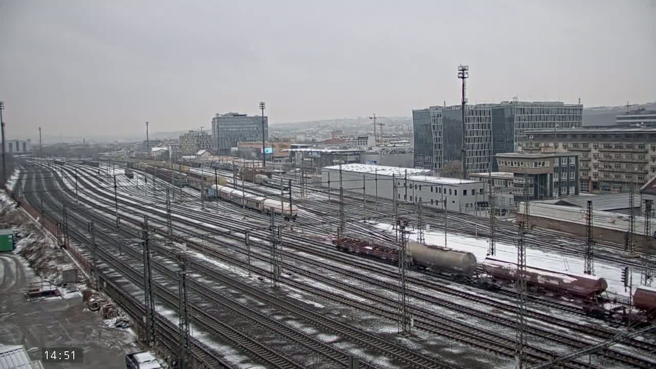 A snow-covered railway yard with numerous train tracks and several freight trains, situated in front of a city skyline under an overcast sky.