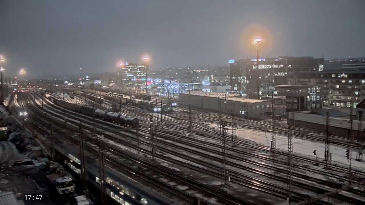 A nighttime view of a snow-covered train yard with multiple tracks and trains, surrounded by illuminated buildings.