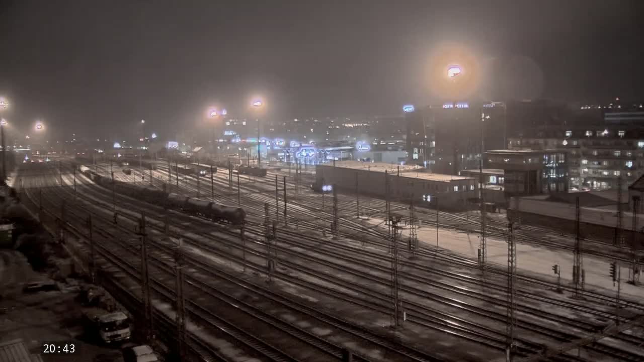 A snow-covered train yard at night is illuminated by numerous lights, with buildings and city lights visible in the background.