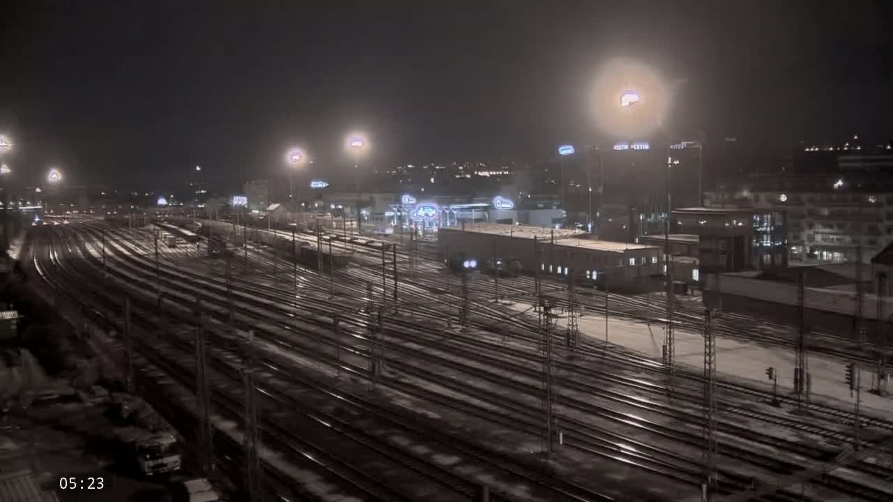 A snow-covered train yard at night, illuminated by numerous lights, with buildings and a city skyline in the background.