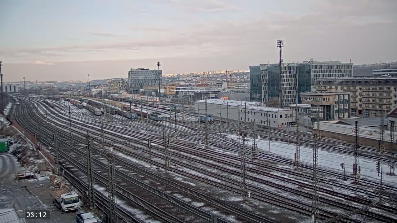 A snow-dusted railway yard with numerous train tracks stretches out before a cityscape of modern and older buildings under a pale sky.