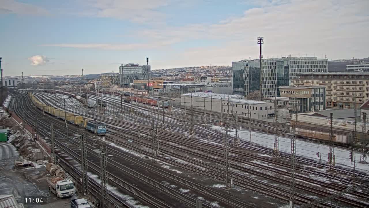 A sprawling railway yard with numerous train cars and tracks, partially covered in snow, sits in front of a city skyline under a cloudy sky.