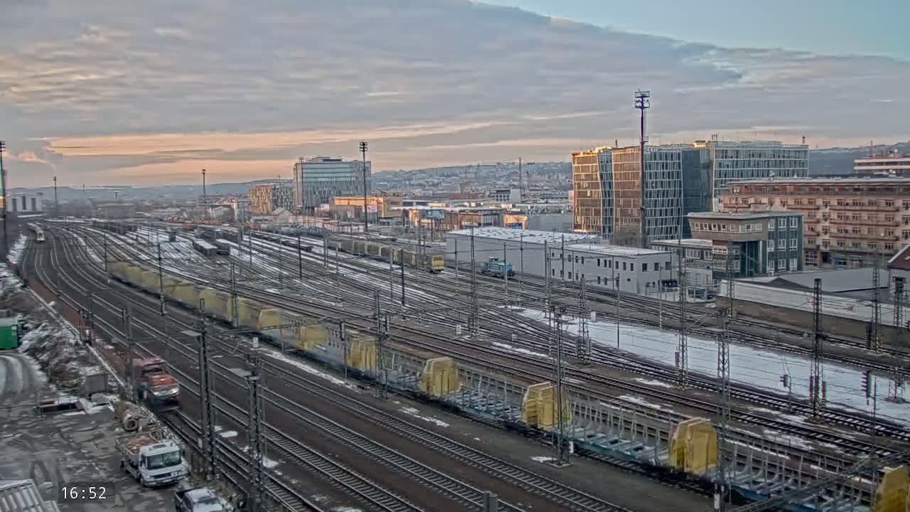 A sprawling rail yard with numerous freight cars and trains under a partly cloudy, sunset sky, with light snow dusting the ground.