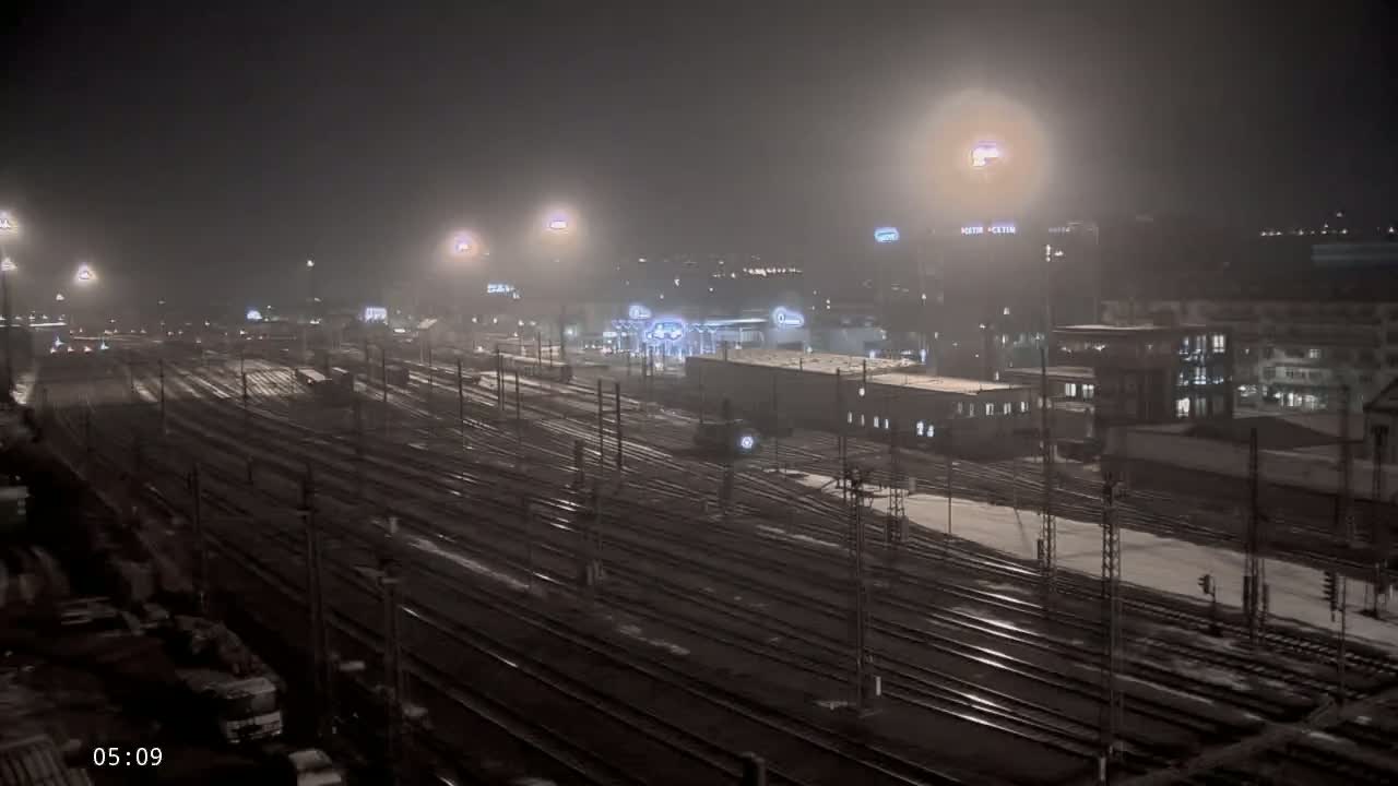 A snowy night scene shows a vast network of train tracks illuminated by artificial lights under a foggy sky.