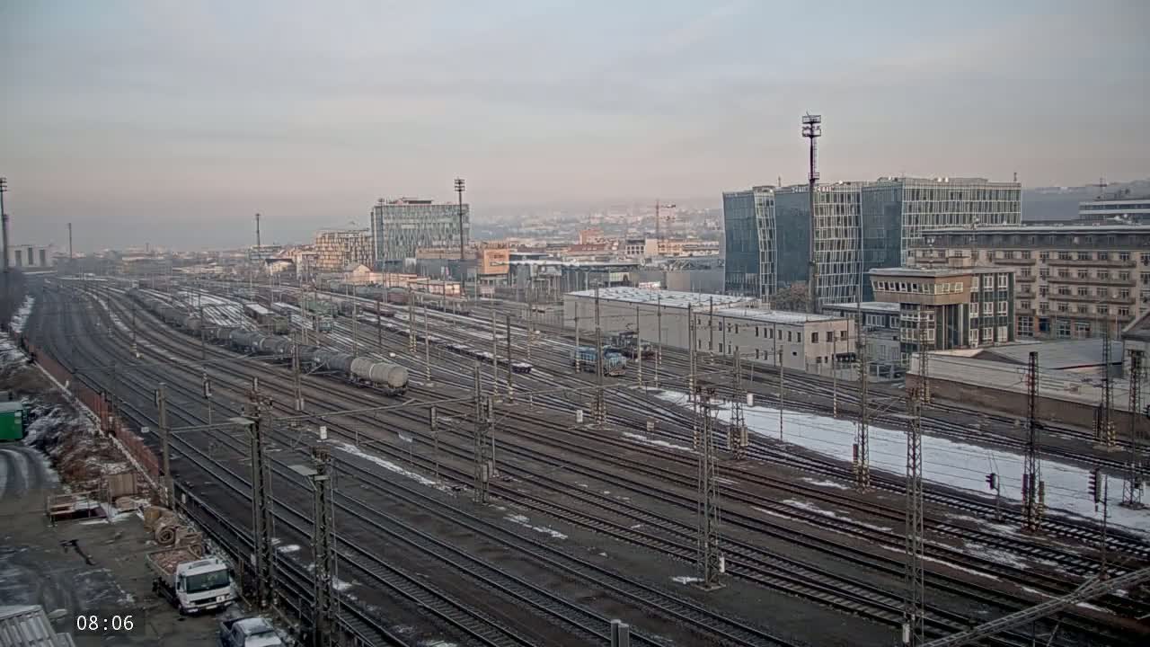 A large railway yard with numerous tracks and freight cars, partially covered in snow, is situated near several modern and older buildings under a cloudy sky.