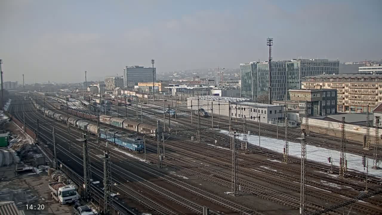 A hazy, sunny day reveals a sprawling railway yard with numerous train cars and tracks, alongside a cityscape of various buildings, some of which have patches of snow on the ground.