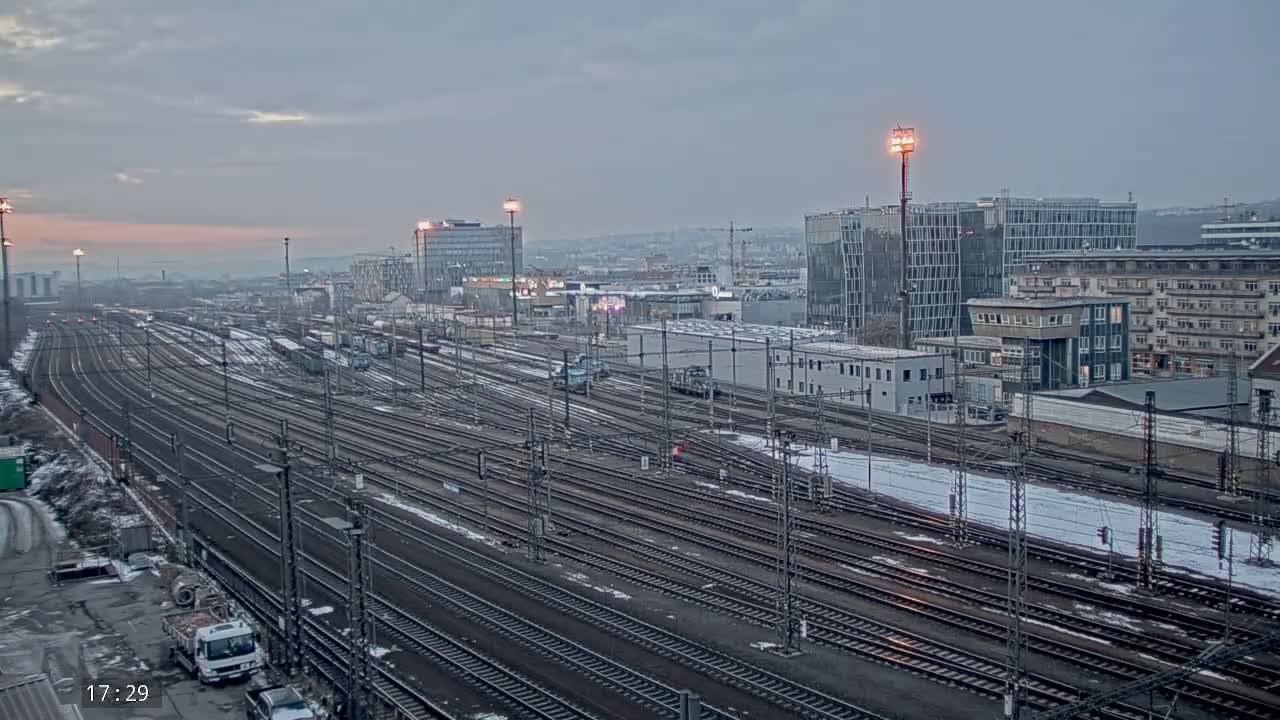 A sprawling railway yard with numerous tracks, partially covered in snow, is situated in front of a city skyline under a dusky, overcast sky.