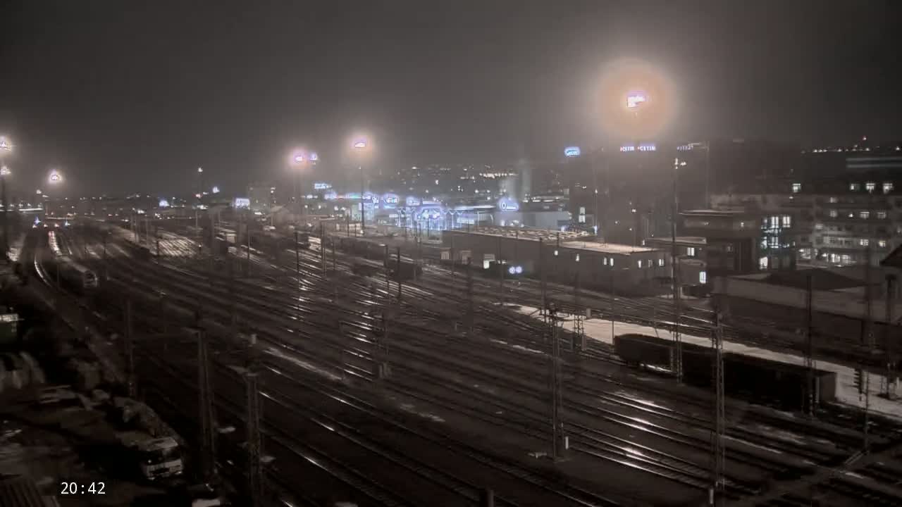 A nighttime view of a snowy, rain-slicked train yard with numerous tracks and buildings in the background under hazy, light-polluted skies.