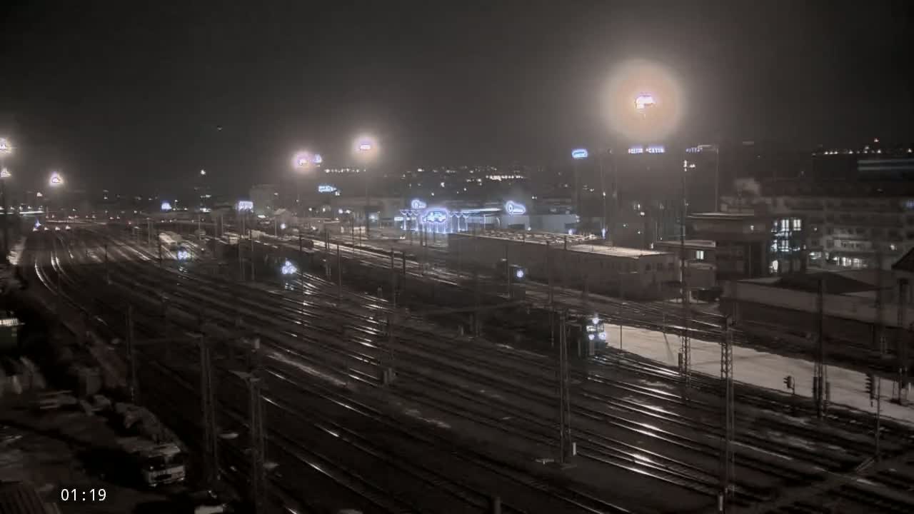 A nighttime view of a snow-covered train yard with multiple tracks and surrounding buildings illuminated by artificial lights.