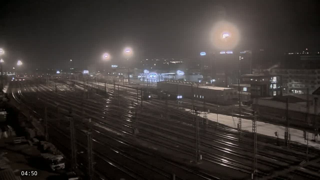 A nighttime view of a snow-dusted railway yard with numerous tracks and buildings under hazy, dimly lit conditions.