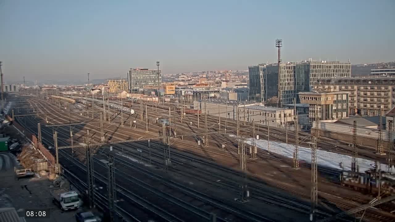 A sprawling railway yard with numerous tracks and train cars is visible under a clear, sunny sky, with some patches of snow on the ground near the tracks and buildings in the background.
