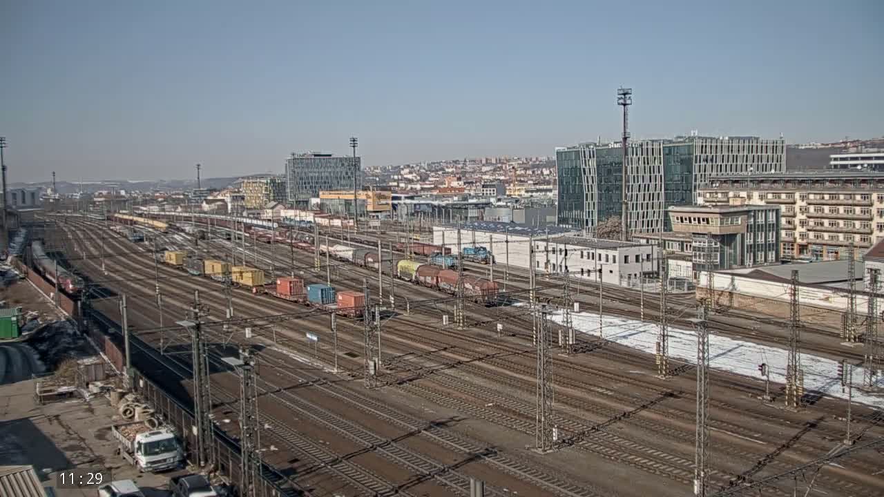 A wide shot of a sprawling railway yard with numerous freight cars and train tracks under a clear, sunny sky, bordered by buildings and patches of snow.