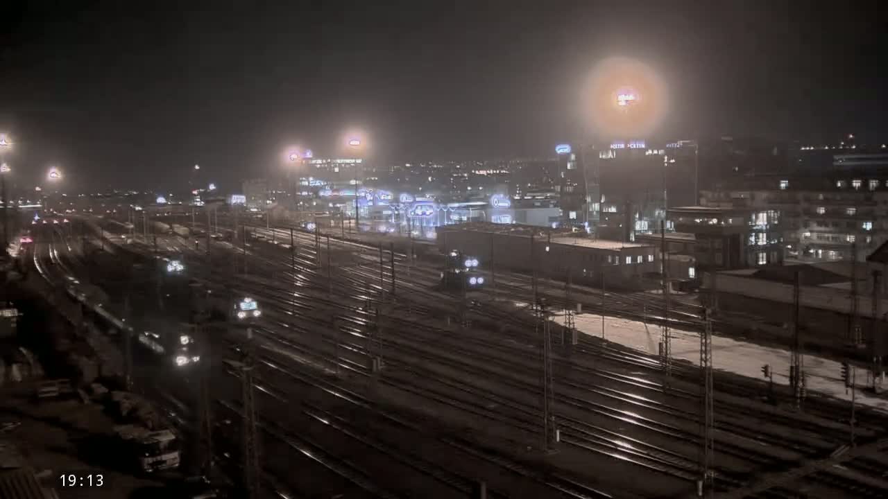 A nighttime view of a train yard with several trains and buildings under light snow.