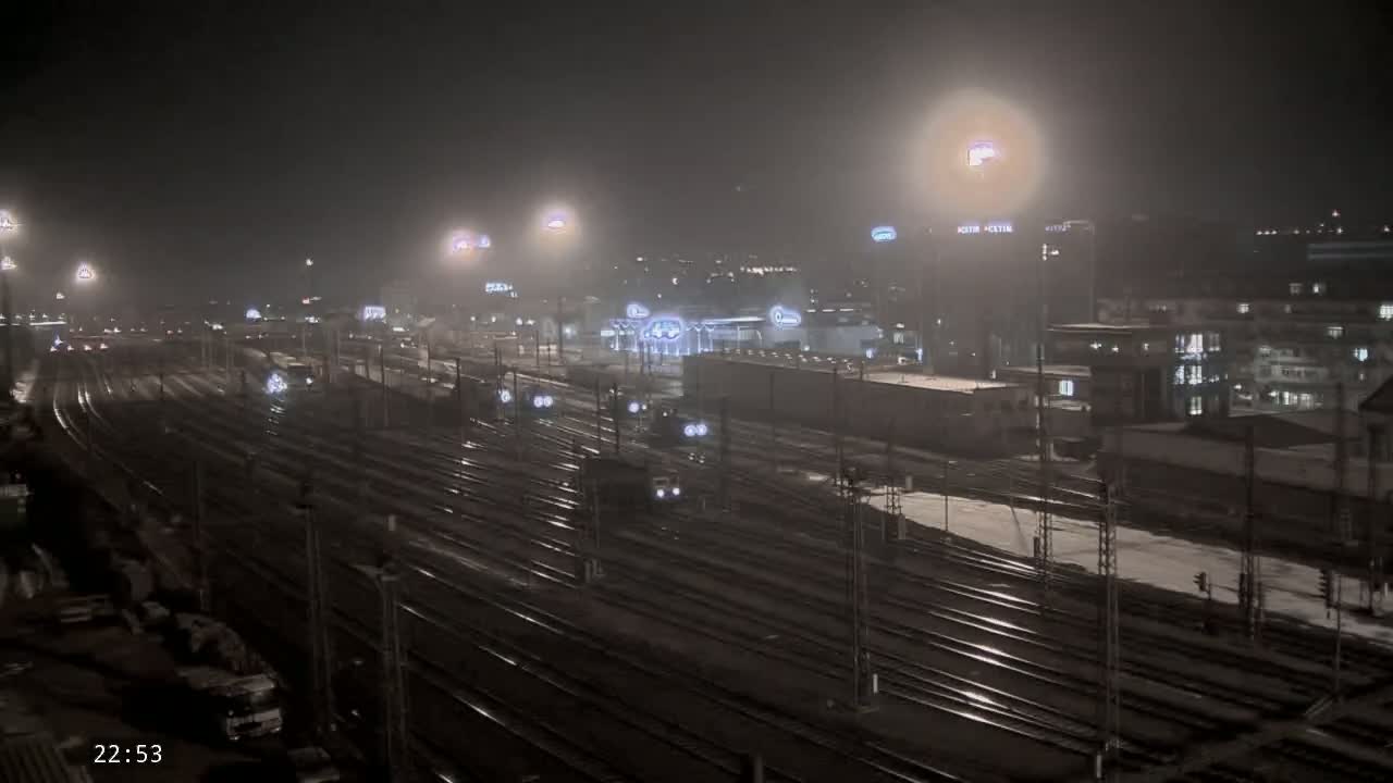 A nighttime view of a snow-covered train yard with multiple sets of tracks and several trains, illuminated by numerous lights.