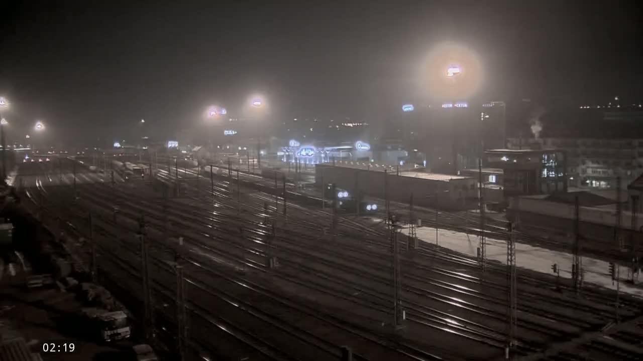 A nighttime view of a snow-covered railway yard with buildings and lights in the background under hazy conditions.