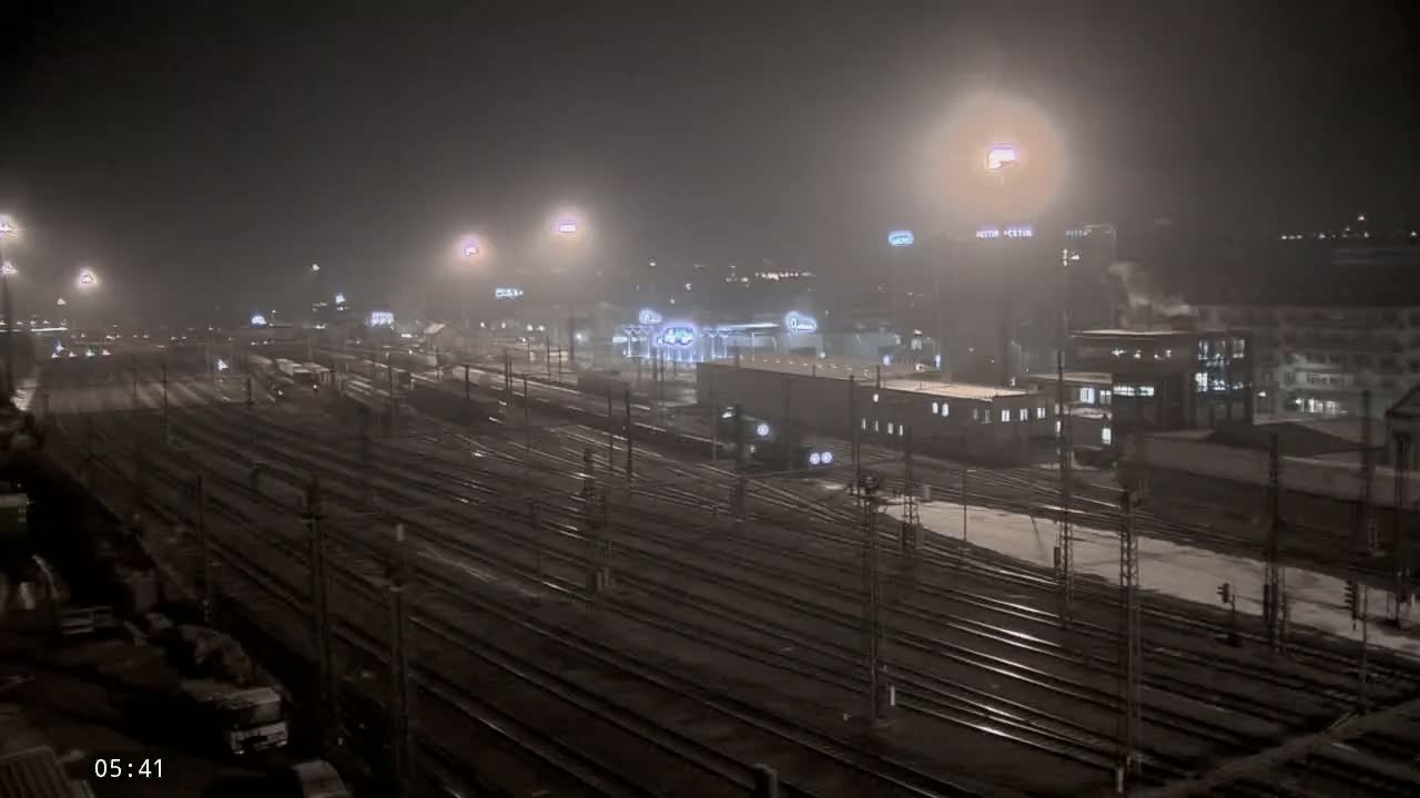 A nighttime view of a snow-covered train yard with numerous tracks and buildings under hazy, light snowfall.