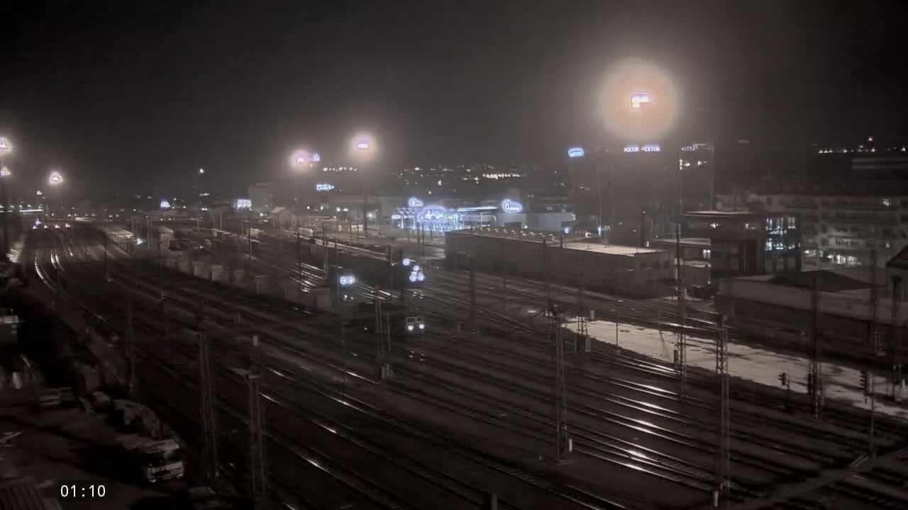A nighttime view of a train yard with several trains and buildings, lightly snow-covered ground, and illuminated by many lights.