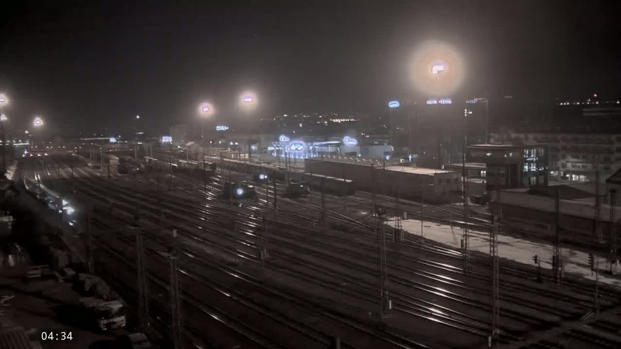 A nighttime view of a snow-dusted railway yard with numerous train tracks, lit by bright lights, and buildings in the background.