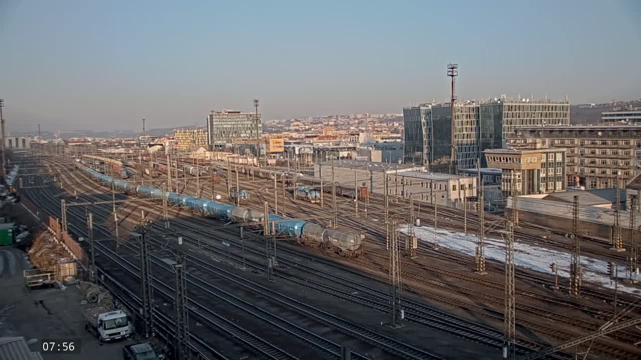 A sprawling rail yard with numerous train cars and tracks, surrounded by buildings on a clear, cold day with patches of snow on the ground.