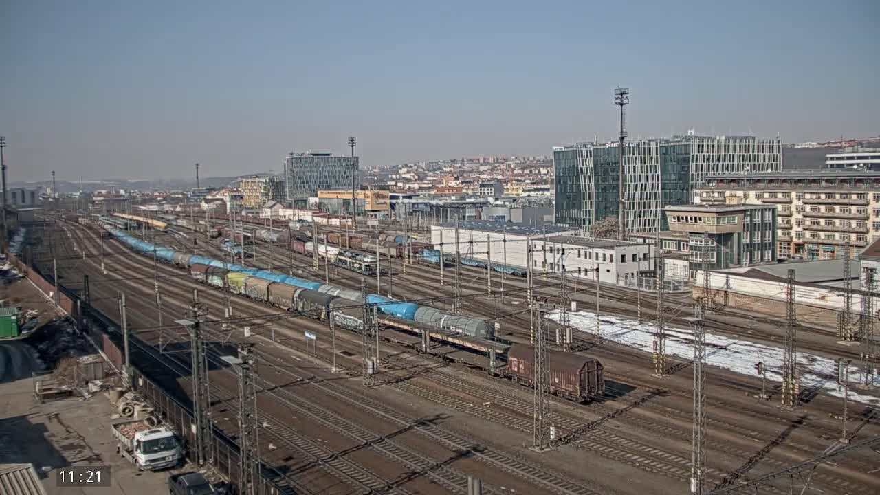 A sprawling railway yard filled with numerous freight trains under a clear, sunny sky, situated beside a city with modern and older buildings.