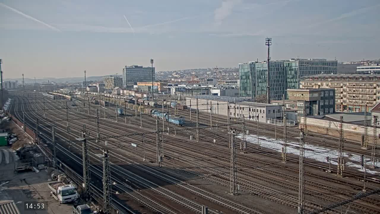 A vast railway yard with numerous tracks and train cars, situated near a city with modern and older buildings under a clear, sunny sky.