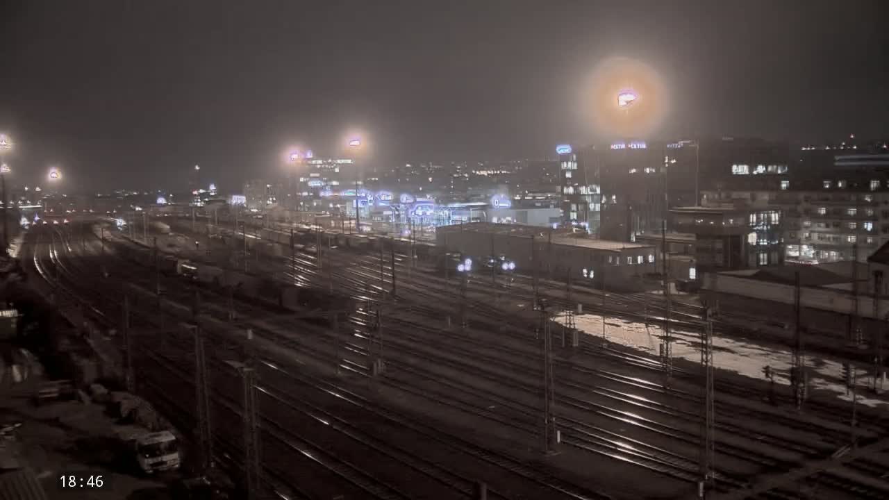 A nighttime view of a train yard and city buildings under a hazy sky with light snow or rain on the ground.