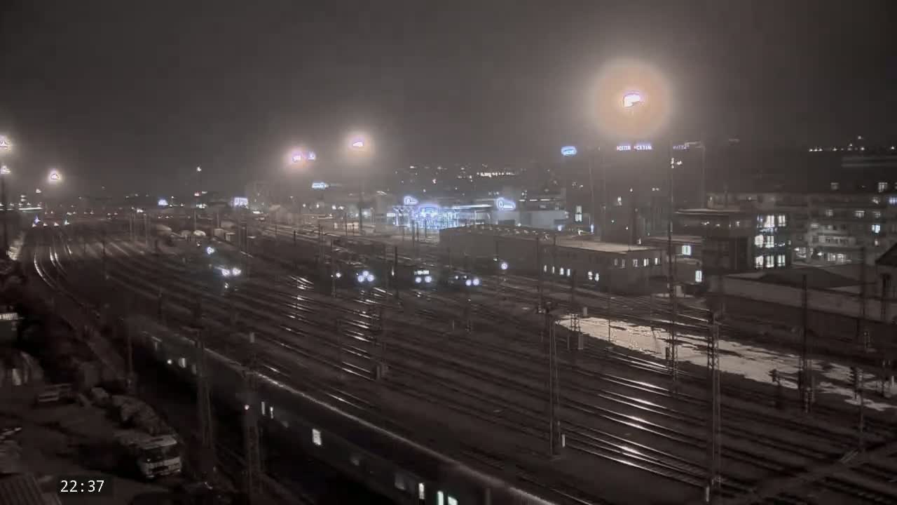 A nighttime view of a train station with numerous train tracks, lit by artificial lights under foggy conditions with some snow or ice on the ground.