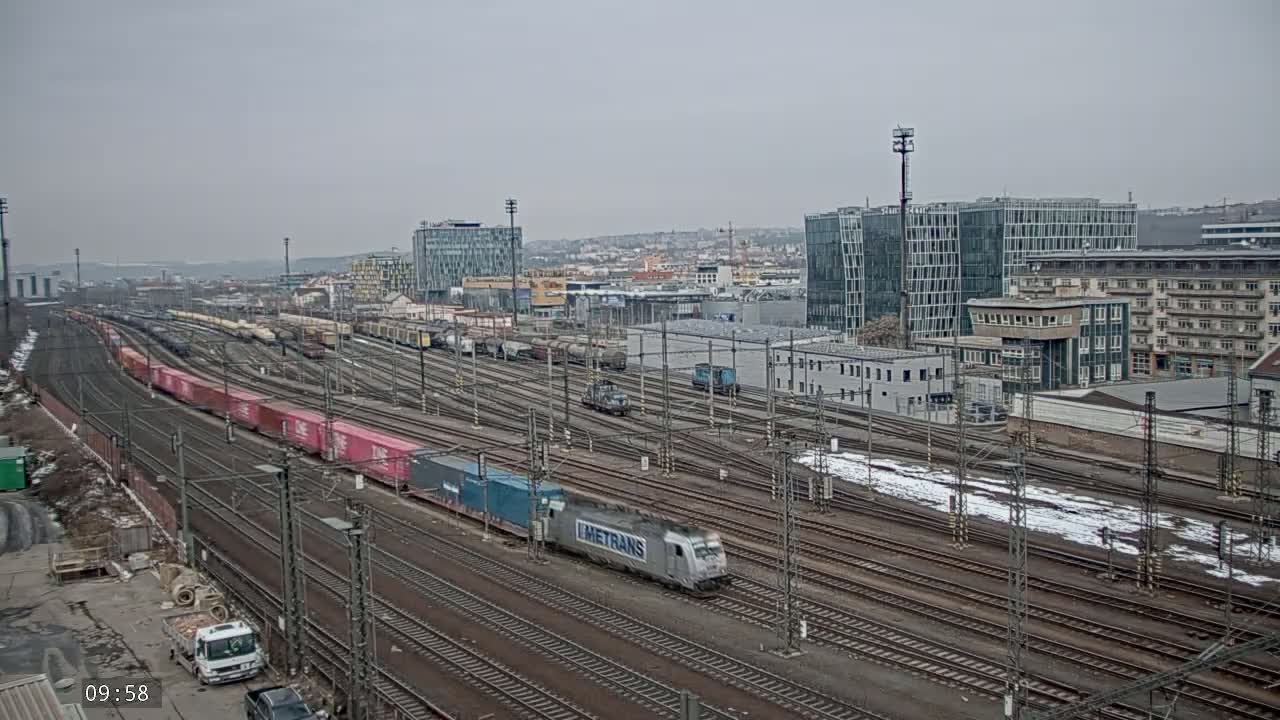 A long freight train carrying shipping containers travels past a railway yard on an overcast day with patches of snow on the ground.