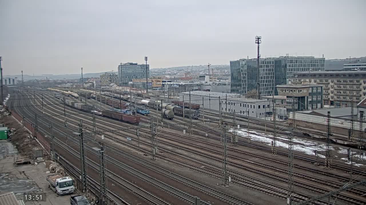 Many railway tracks and freight trains are visible in a sprawling railway yard, with overcast skies and patches of snow on the ground in the background are modern and older buildings.