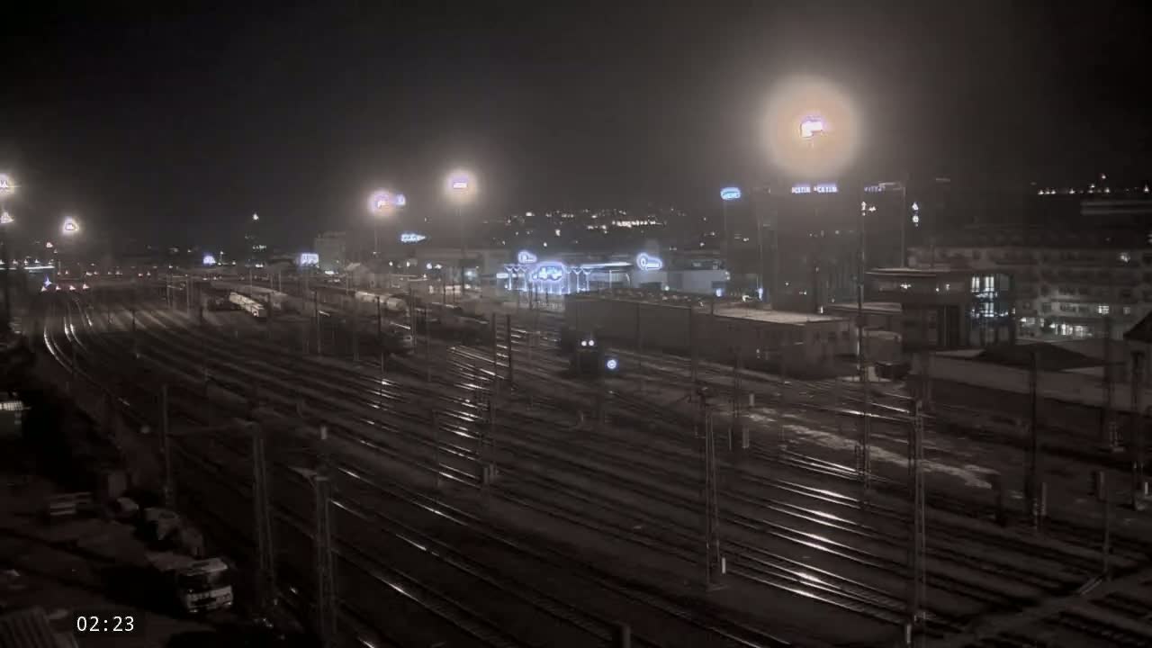 A nighttime view of a train yard with many railway tracks, illuminated by artificial lights, with some buildings and vehicles visible in the background.