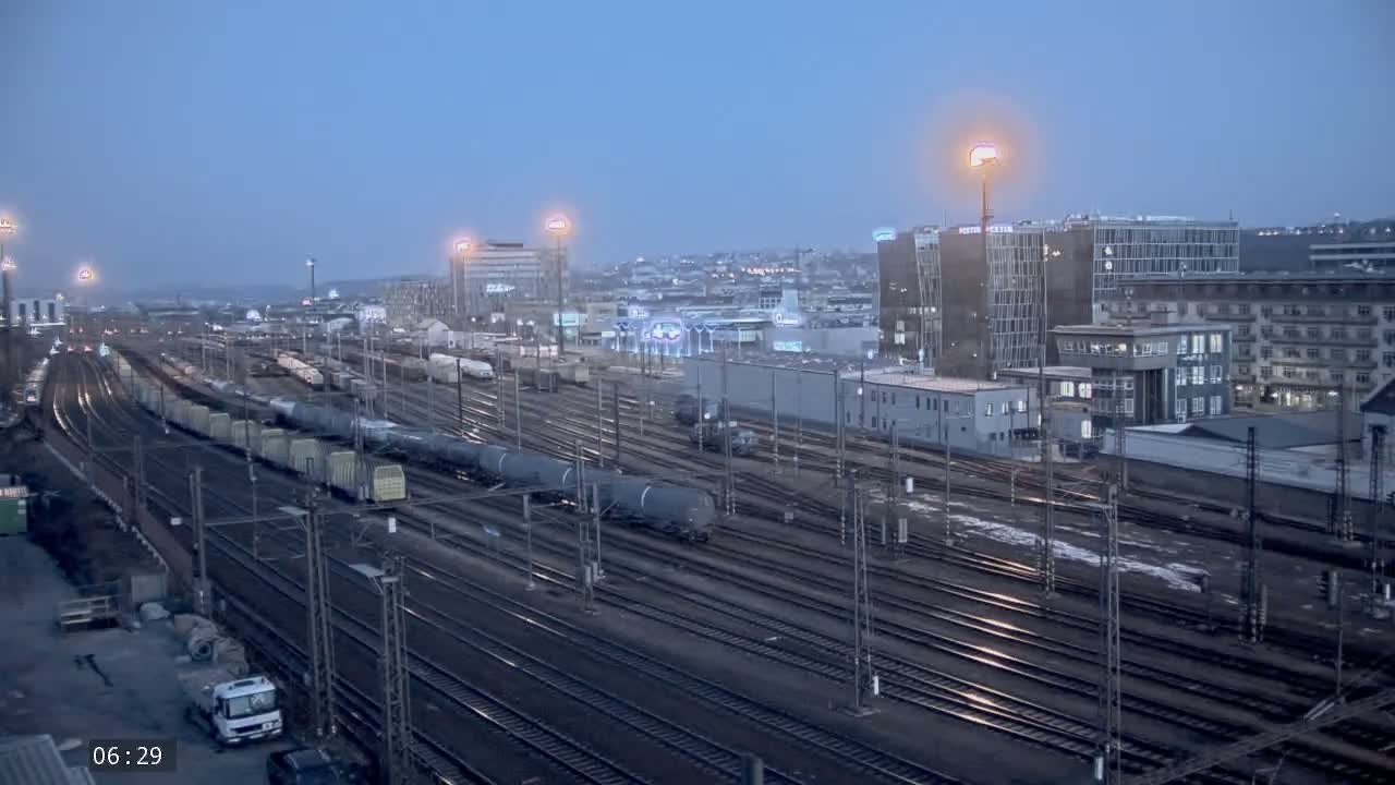 Under a twilight sky, numerous train cars sit on a sprawling network of train tracks in a city with several multi-story buildings.