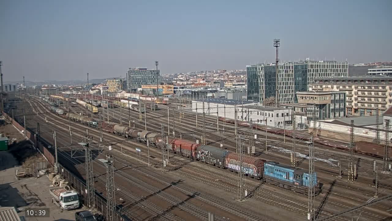 A vast railway yard filled with numerous freight trains and cars under a clear, sunny sky, adjacent to a city with modern and older buildings.