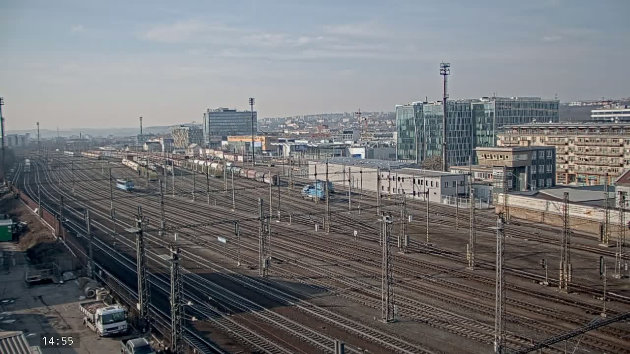 A vast railway yard with numerous tracks and parked trains is situated in front of a city skyline under a mostly clear sky.