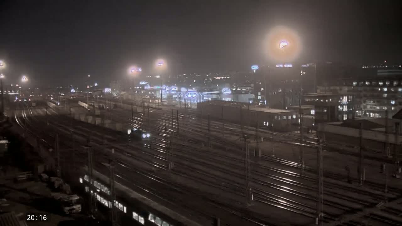 A nighttime view of a train yard with numerous train tracks, lit buildings in the background, and hazy atmospheric conditions.